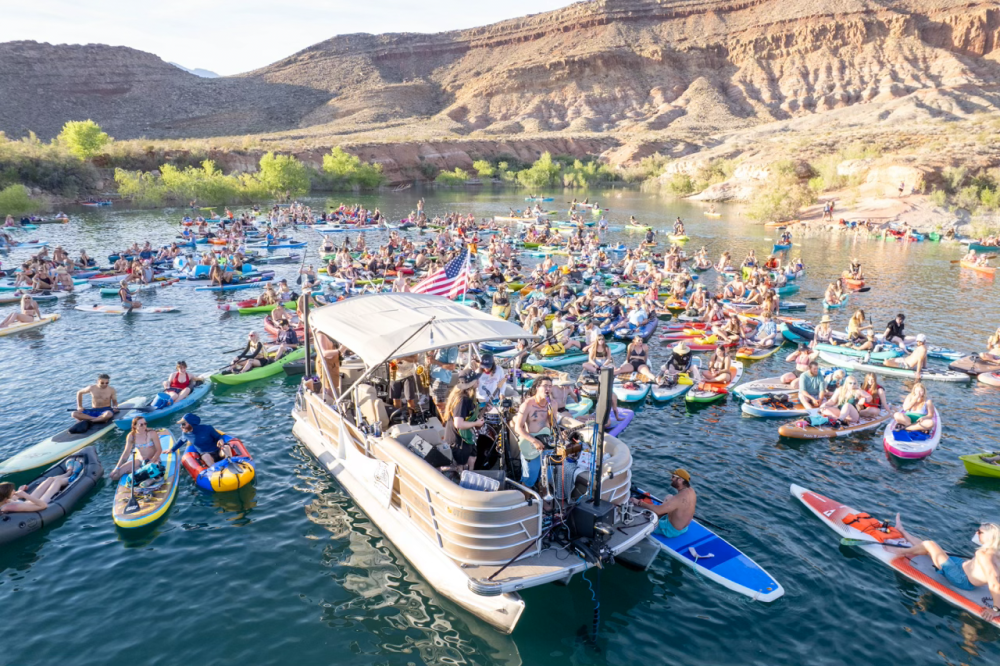 Crowd of people on kayaks and paddleboards near a boat on a lake with rocky hills in the background.