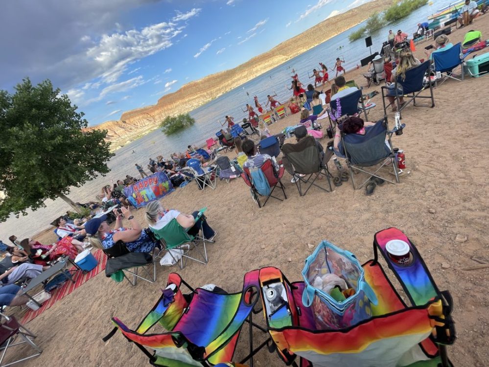 Crowd seated on beach chairs watching performers by a lake under a blue sky.